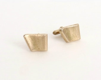 May include: A pair of gold-toned cufflinks featuring a geometric, trapezoidal shape with a brushed, matte texture. Each cufflink has a flat, angled front surface and a small metal post extending from the back for attachment to a shirt cuff.
