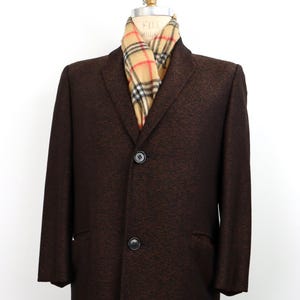 May include: A brown herringbone-patterned overcoat with a notched collar and two-button closure. A plaid scarf in tan, red, and black is draped around the neck. The coat is displayed on a mannequin.