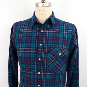 Peut inclure: Chemise à carreaux bleu sarcelle, marine et bordeaux à manches longues, présentée sur un mannequin. Elle comporte une fermeture boutonnée, une poche poitrine et des boutons nacrés. Le motif à carreaux est un design classique.