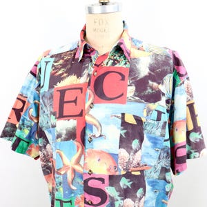 May include: Short-sleeved button-down shirt with a colorful, all-over print featuring marine life and large block letters. The shirt has a collared neckline and a single chest pocket. The color palette includes red, blue, green, and purple.