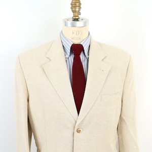 Może przedstawiać: Beżowy blazer z lnu z zapięciem na trzy guziki i klapami kieszeni. Blazer jest na manekinie, który nosi niebiesko-białą koszulę w paski i bordowy krawat.