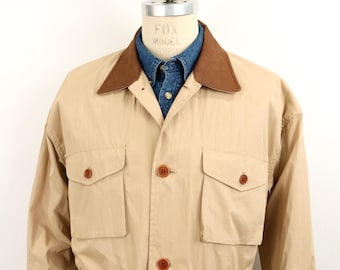 May include: A tan field jacket featuring a brown corduroy collar, four front flap pockets with button closures, and a central button-down placket. The jacket is layered over a blue denim button-down shirt, visible at the collar and cuffs.