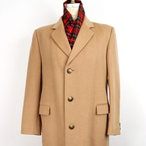 May include: A tan overcoat with a classic design. The coat features a notched collar, three buttons, and two front pockets. A red, black, and green plaid scarf is visible at the neck. The coat is displayed on a mannequin.
