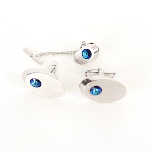 May include: A set of silver cufflinks and a tie tack with blue gemstone accents. The cufflinks and tie tack are oval shaped.