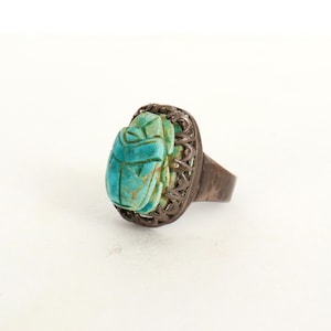 May include: A ring featuring a carved turquoise-colored scarab beetle centerpiece set within a dark, oxidized metal frame. The band is smooth and dark, contrasting with the textured, light blue-green stone beetle that sits prominently on top.