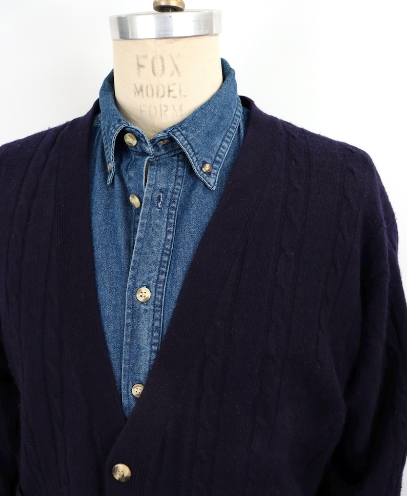 Puede incluir: Un c&aacute;rdigan azul marino de punto trenzado con cuello en V, usado sobre una camisa vaquera abotonada. El c&aacute;rdigan tiene tres botones visibles. La camisa tiene cuello abotonado y pu&ntilde;os abotonados. La cabeza del maniqu&iacute; es visible.
