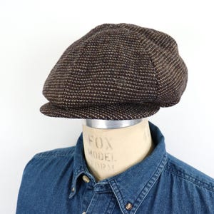 May include: A brown tweed newsboy cap with a textured pattern. The cap is displayed on a mannequin head over a denim shirt. The cap has a rounded crown and a short, stiff brim.