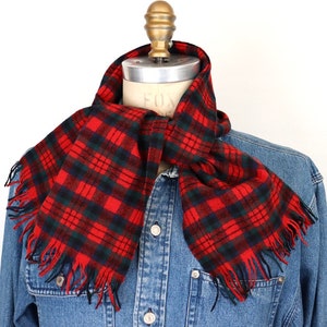 Pode incluir: Cachecol xadrez vermelho, verde e preto com franjas. O cachecol está sobre uma jaqueta jeans em um manequim. O padrão xadrez é um design clássico, adequado para o clima frio.