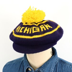 May include: A dark blue knit hat with a yellow pom-pom on top. The hat features a yellow horizontal stripe and the word MICHIGAN knitted in yellow block letters across the front. It has a short, stiff brim and a rounded, soft crown structure.