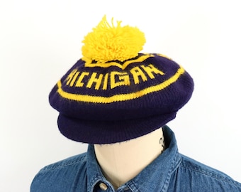 May include: A dark blue knit hat with a yellow pom-pom on top. The hat features a yellow horizontal stripe and the word MICHIGAN knitted in yellow block letters across the front. It has a short, stiff brim and a rounded, soft crown structure.
