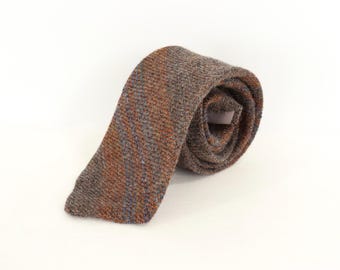 May include: A rolled necktie featuring a textured, woven fabric in shades of brown, gray, and rust orange. The tie displays a subtle diagonal striped pattern throughout its length, with a visible white fabric label tucked inside the rolled coil.