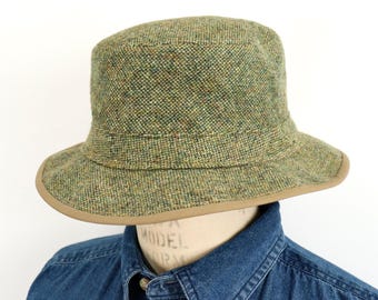 May include: A bucket hat crafted from a textured, multi-colored green and brown tweed fabric. The hat features a structured crown and a downward-sloping brim finished with a smooth, solid tan-colored trim along the outer edge of the brim.
