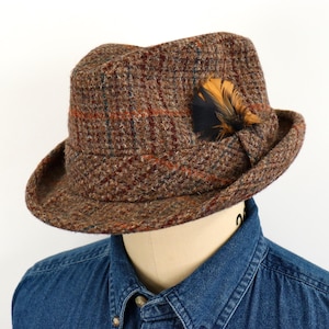 May include: A brown tweed fedora hat featuring a textured plaid pattern with subtle blue and orange lines. The hat is adorned with a decorative dark and golden-brown feather tucked into the side band, showcasing a structured crown and brim.