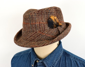 May include: A brown tweed fedora hat featuring a textured plaid pattern with subtle blue and orange lines. The hat is adorned with a decorative dark and golden-brown feather tucked into the side band, showcasing a structured crown and brim.