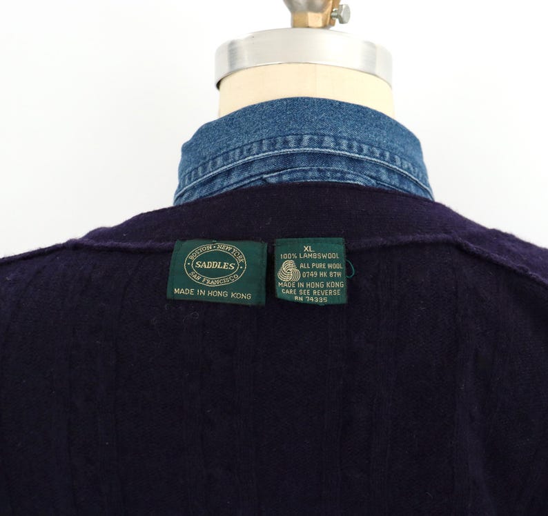 Puede incluir: Primer plano de un su&eacute;ter de lana azul marino oscuro con cuello de camisa de mezclilla. El su&eacute;ter tiene dos etiquetas verdes con texto blanco. Una etiqueta dice "Saddles San Francisco Made in Hong Kong". La otra dice "XL 100% Lambswool All Pure Wool".