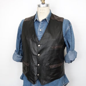 Puede incluir: Un chaleco de cuero negro con cuello y bolsillos de ante marrón. El chaleco tiene cierre de botones y se lleva sobre una camisa vaquera azul.