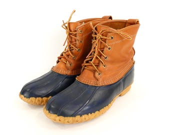 May include: A pair of ankle-height duck boots featuring tan leather uppers with a lace-up closure and brass eyelets. The lower portion is constructed from dark navy blue rubber with a textured toe cap and a light tan lugged rubber sole.