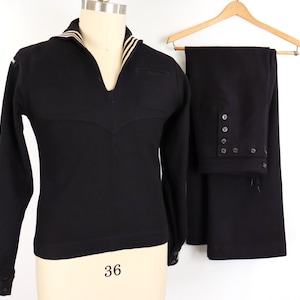May include: A black long-sleeve pullover shirt with a V-neck collar featuring white stripes, paired with matching black trousers that have a button-front closure. The shirt has white striped cuffs and the trousers are displayed on a wooden hanger.