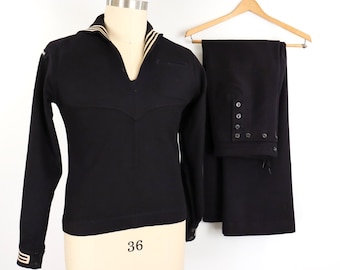 May include: A black long-sleeve pullover shirt with a V-neck collar featuring white stripes, paired with matching black trousers that have a button-front closure. The shirt has white striped cuffs and the trousers are displayed on a wooden hanger.