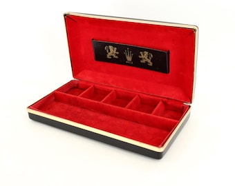 May include: A rectangular black jewelry box with a hinged lid and gold-tone trim. The interior is lined with bright red velvet and features five small compartments. A black plaque inside the lid displays a gold crown logo flanked by two lions.