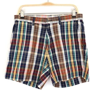 Può includere: Pantaloncini a quadri multicolore, con quadri blu navy, rossi, verdi e gialli, appesi a una gruccia di legno. I pantaloncini hanno una chiusura con bottoni e passanti per cintura. I pantaloncini sono realizzati in tessuto leggero.