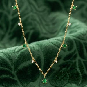 Puede incluir: Un collar de cadena dorada con pequeñas piedras preciosas verdes de corte cuadrado que alternan con piedras blancas redondas y transparentes, terminado con un colgante de piedra verde en forma de pera en el centro, sobre una fina cadena de eslabones.