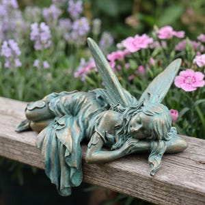 May include: A teal and bronze-toned fairy figurine in a reclining position with wings extended upward. The figure has long hair, closed eyes, and is wearing a dress with ruffled details, resting its head on one arm while lying on a wooden surface.