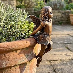 Peut inclure: Une figurine de fée couleur bronze avec des ailes détaillées et une robe courte, conçue pour se percher sur le bord d'un pot en terre cuite. La silhouette présente des cheveux ondulés et une main posée sur le rebord du pot.