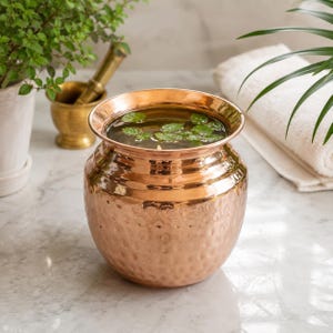 May include: A round, hammered copper vessel with a wide, flared rim. The container is filled with clear liquid and several small green leaves floating on the surface. The metal surface has a polished, reflective finish with a textured pattern.