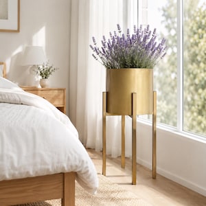 Op de afbeelding: Een cilindervormige gouden metalen plantenbak op een hoge, vierpotige gouden metalen standaard. De plantenbak is gevuld met een dicht arrangement van paarse lavendelbloemen. De structuur heeft een gladde, metalen afwerking en verticaal ontwerp.