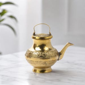 Puede incluir: Un recipiente de latón pulido con cuerpo redondeado, cuello estrecho y pico curvo. El exterior está decorado con intrincados patrones florales en relieve y bandas horizontales. Un asa de metal delgada y arqueada está unida a la tapa.