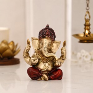 May include: A brass-toned Ganesha figurine in a seated position with four arms, featuring a dark red crown and matching red dhoti. The deity has an elephant head with a trunk curved to the left, large ears, and a prominent Om symbol on the forehead.