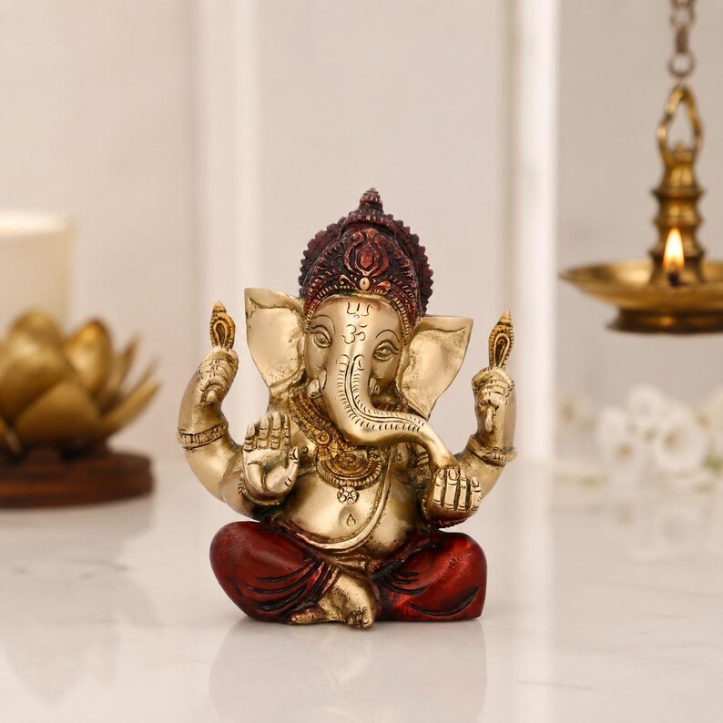 May include: A brass-toned Ganesha figurine in a seated position with four arms, featuring a dark red crown and matching red dhoti. The deity has an elephant head with a trunk curved to the left, large ears, and a prominent Om symbol on the forehead.