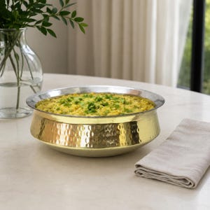 May include: A round, hammered brass serving bowl with a silver-toned rim, filled with a yellow grain-based dish topped with fresh green herbs. The bowl features a textured, dimpled exterior surface and a wide, shallow shape for serving food.