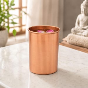 Könnte beinhalten: Ein zylindrischer Kupferbecher mit einer glatten, metallischen Oberfläche. Das Gefäß ist mit Flüssigkeit gefüllt und zeigt ein einzelnes rosa Blütenblatt, das auf der Oberfläche schwimmt, was eine schlichte Ästhetik für diesen Trinkbecher schafft.