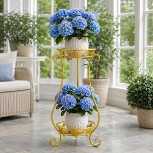 Op de afbeelding: Een plantenstandaard met twee niveaus van goudkleurig metaal met gebogen poten en ronde planken. Elke plank bevat een witte, getextureerde keramische pot met een levendige blauwe hortensia met groene bladeren.