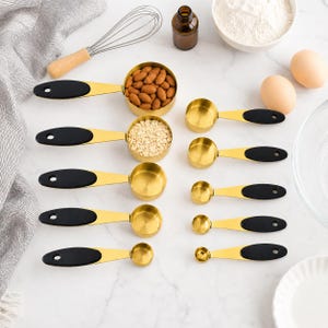 Peut inclure: Un ensemble de dix tasses et cuillères à mesurer en acier inoxydable doré avec des poignées en silicone noir. Le set comprend différentes tailles pour ingrédients secs et liquides, disposées en deux colonnes sur une surface blanche.