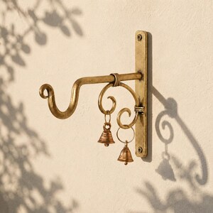 Puede incluir: Un gancho de pared de latón con un brazo curvo y una placa de montaje vertical fijada con dos tornillos. Dos pequeñas campanas decorativas de latón cuelgan de adornos metálicos en forma de voluta unidos al brazo de soporte principal.
