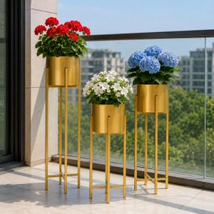 Op de afbeelding: Drie goudkleurige metalen plantenstandaards van verschillende hoogtes, elk met een cilindrische pot in een frame met vier poten. De standaards bevatten levendige potbloemen: rode geraniums, witte bloesems en blauwe hortensia's op een rij.