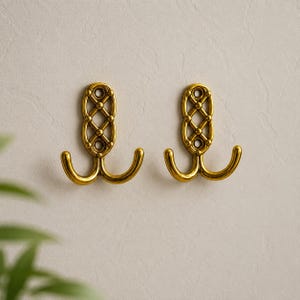 以下が含まれることがあります： 金色の金属製壁掛けフック2つ。垂直の取り付けプレートには装飾的な格子模様が施されています。各フックには外側に伸びる2本の湾曲したアームがあり、プレートの上下にはネジ用の円形の穴が開いています。