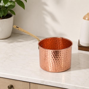 May include: A round, hammered copper saucepan with a long, polished brass handle. The pot features a distinct metallic texture across its exterior surface and a small pouring spout on the rim, showcasing a warm, reflective finish throughout.