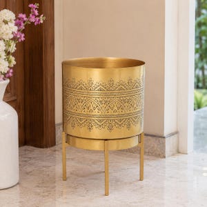 May include: A cylindrical gold-colored metal planter featuring a detailed black etched pattern around the upper section. The planter is supported by a matching gold-colored metal stand with four thin legs and a circular base ring for stability.