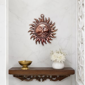 May include: A copper-coloured metal sun wall hanging featuring a central face with a moustache and a forehead mark, surrounded by stylised flame-like rays. The circular piece includes a small loop at the top for mounting on a vertical surface.