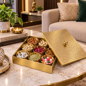 Puede incluir: Una caja cuadrada de metal dorado con tapa extraíble que presenta un intrincado patrón floral y un pomo central. La caja contiene nueve cuencos redondos dorados pequeños llenos de varios frutos secos, semillas y caramelos de colores.