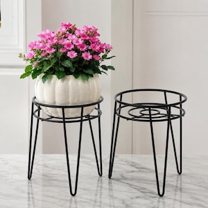 Op de afbeelding: Twee zwarte metalen plantenstandaards met haarspeldpoten en ronde frames. Eén standaard bevat een witte, geribbelde, ronde keramische pot met een bosje kleine roze bloemen en groene bladeren, terwijl de andere standaard leeg is.