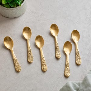 Peut inclure: Un ensemble de six cuillères en métal doré disposées en groupe. Chaque cuillère présente un bol lisse et arrondi ainsi qu'un manche orné d'un motif en relief texturé de feuilles et de grille s'étendant du col jusqu'à la base du manche.