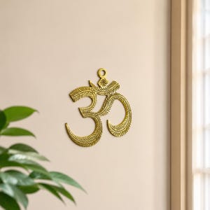 Könnte beinhalten: Ein goldfarbenes Wandornament mit dem Om-Symbol. Das Design weist eine blattartige Textur auf der gesamten Oberfläche auf und enthält eine kleine kreisförmige Öse an der Oberseite zum Aufhängen. Das Objekt ist flach und metallisch.
