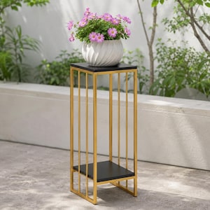Op de afbeelding: Een hoge, rechthoekige plantenstandaard met een goudkleurig metalen frame met verticale stangen, een zwart vierkant bovenoppervlak en een bijpassende zwarte onderste plank. Een witte, gedraaide keramische pot met roze bloemen.