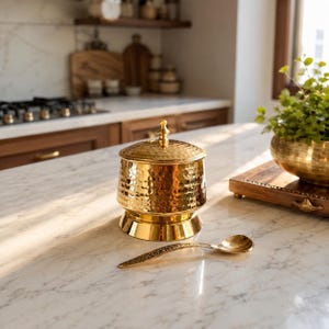 Puede incluir: Un recipiente de metal martillado de color dorado con una tapa a juego que presenta un pequeño pomo, acompañado de una cuchara delgada y a juego de color dorado con mango texturizado, sobre una superficie blanca con vetas grises.