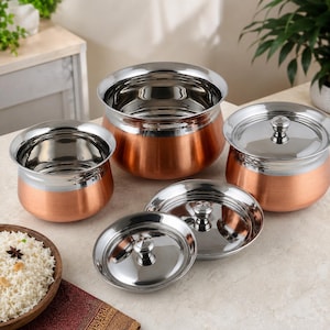 May include: A set of three round cooking pots with copper-toned exterior bases and polished stainless steel interiors and rims. Two of the pots have matching stainless steel lids with central knobs, while one pot remains open and uncovered.